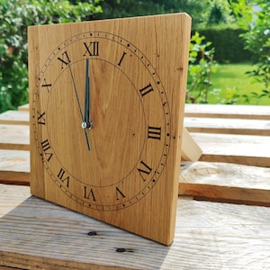 May include: A square wooden clock with Roman numerals and black hands. The clock is made of light-colored wood and has a stand on the back. The clock face displays the time as nearly 12 o'clock.