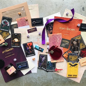 Könnte beinhalten: Ein Brettspiel-Set mit einem lila und goldenen Spielführer, einem roten Samtbeutel, einem gelben Kartenspiel und mehreren schwarzen und goldenen Karten mit Illustrationen. Das Spiel heißt "The Last Afterwards" und "Secret Histories".