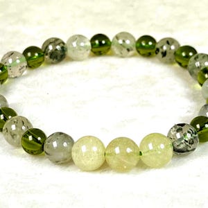 Puede incluir: Una pulsera elástica compuesta por cuentas redondas alternas en tonos verde oliva, transparente claro con inclusiones oscuras y amarillo pálido. Las cuentas están dispuestas en un patrón circular, creando un bucle continuo de esferas de piedra pulida.