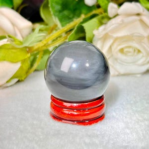 May include: A smooth, polished dark gray sphere with subtle, lighter gray horizontal banding, resting on a circular, glossy reddish-brown wooden stand. The sphere exhibits a reflective surface with a distinct sheen, showcasing natural mineral patterns.