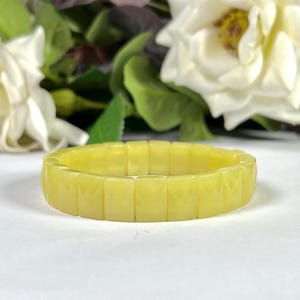 Peut inclure: Un bracelet jaune clair avec un design rectangulaire. Le bracelet est composé de blocs individuels et facettés, créant un aspect texturé. Des roses blanches et du feuillage vert sont en arrière-plan.