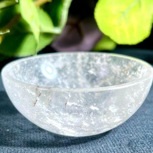 Peut inclure: Un petit bol semi-circulaire taillé dans un cristal de quartz transparent et translucide. Le matériau présente des fractures internes naturelles, des inclusions et une surface polie et lisse qui reflète la lumière sur sa structure ouverte.