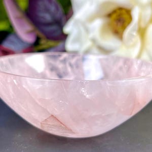 Puede incluir: Un pequeño cuenco de cuarzo rosa claro. El cuenco tiene una superficie lisa y pulida y una forma redondeada. El cuenco se coloca sobre un fondo borroso de flores blancas y moradas. Ideal para guardar objetos pequeños.