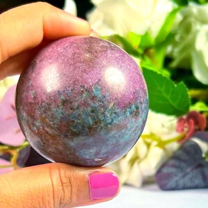 Può includere: Una gemma sferica lucidata, che mostra una miscela di colori viola, blu e bianco. La pietra è tenuta in mano. Lo sfondo presenta fiori bianchi e foglie verdi.