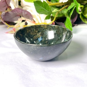 Pode incluir: Uma pequena tigela redonda com uma superfície escura e manchada. A tigela tem um exterior liso e polido e um interior ligeiramente iridescente com toques de azul e dourado. A tigela é provavelmente feita de pedra ou material semelhante.