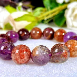 May include: A beaded bracelet featuring polished, spherical gemstones in shades of deep purple, translucent orange, and reddish-brown. The beads are arranged in a circular pattern, showcasing natural inclusions and variations in color and clarity.
