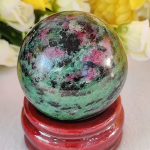 Könnte beinhalten: Eine grüne, schwarze und rosafarbene Edelsteinkugel auf einem roten Holzständer. Die Kugel ist poliert und hat eine glatte Oberfläche.