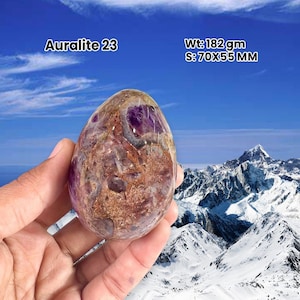 May include: Auralite 23 gemstone, held in a hand, weighing 182 grams and measuring 70x55 mm. The stone displays a blend of purple, brown, and clear hues. The backdrop features a mountain range under a blue sky.