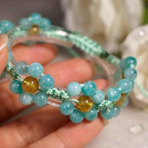 May include: A bracelet featuring a clear circular base adorned with light blue and amber-colored beads arranged in flower-like patterns, connected by light green woven cord, with a single bead at the end of a dangling string.