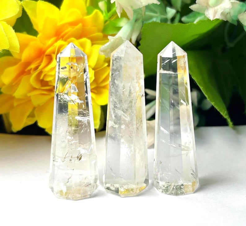 May include: Three clear quartz crystal points with a faceted design. The crystals are displayed against a backdrop of yellow and green flowers. The crystals have a natural, translucent appearance.