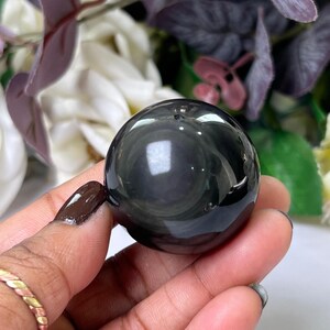 Può includere: Una sfera lucida di ossidiana nera. La sfera ha una superficie riflettente con motivi a spirale ed è tenuta in una mano. Lo sfondo presenta fiori sfocati nelle tonalità del viola, bianco e verde.