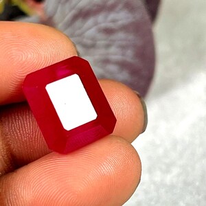 May include: A faceted, rectangular ruby gemstone with a white center. The gem is held between fingers, showcasing its deep red color and geometric shape. The gem's edges are precisely cut, reflecting light.