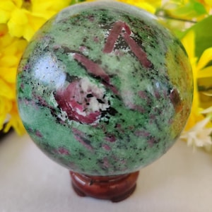 Könnte beinhalten: Eine grüne, rosafarbene und schwarze Edelsteinkugel auf einem Holzständer. Die Kugel ist poliert und hat ein wirbelndes Muster.