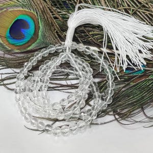 May include: A clear crystal beaded necklace with a white tassel. The beads are round and faceted, reflecting light. The necklace is displayed against a backdrop of peacock feathers, with shades of green, brown, and blue.