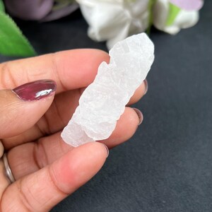 Puede incluir: Un cristal de cuarzo blanco en bruto se sostiene en una mano. El cristal tiene una superficie rugosa y texturizada y una forma irregular. El fondo es de color oscuro, con toques de flores moradas y blancas.