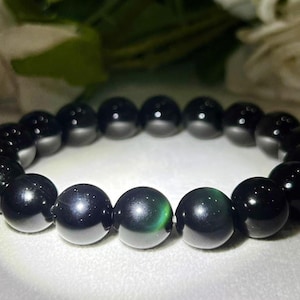 Könnte beinhalten: Ein elastisches Armband bestehend aus glatten, runden, polierten schwarzen Perlen. Die Perlen weisen einen subtilen, reflektierenden Glanz mit schwachen grünen Lichtreflexen auf ihren Oberflächen auf und sind in einer kreisförmigen Schleife angeordnet.