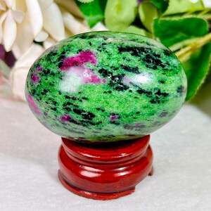 May include: A polished, oval-shaped zoisite stone featuring a vibrant green base with prominent inclusions of deep magenta ruby and dark black hornblende, resting securely on a circular, dark reddish-brown wooden display stand.