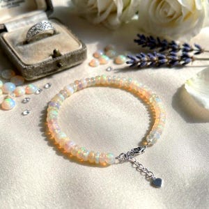 May include: A beaded bracelet featuring round, translucent opal beads with iridescent flashes of color. The bracelet includes a silver-toned metal lobster claw clasp, an adjustable chain extension, and a small heart-shaped charm at the end.