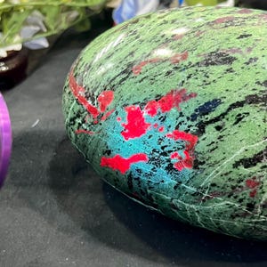 以下が含まれることがあります： 緑色のベースカラーに黒い斑点、鮮やかな赤とターコイズのパッチがアクセントになった、磨かれた楕円形の石。石の表面は滑らかで反射性があり、自然で有機的な模様を示しています。