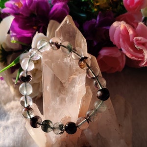 May include: A bracelet made of clear, green, and black beads. The bracelet is resting on a large, clear quartz crystal.