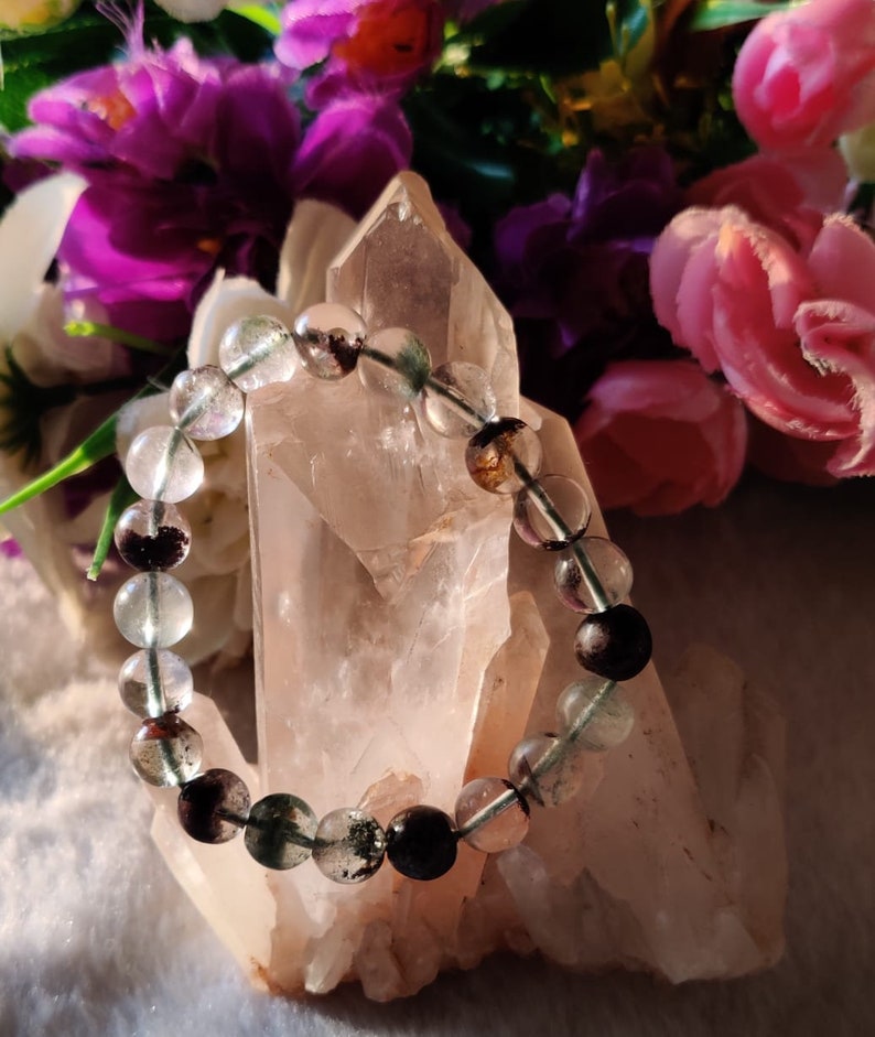 May include: A bracelet made of clear, green, and black beads. The bracelet is resting on a large, clear quartz crystal.