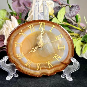 Puede incluir: Un reloj decorativo de rodaja de ágata con números romanos y manecillas doradas. La esfera del reloj es una mezcla de marrón, beige y blanco, colocada en un soporte de acrílico transparente. Un artículo de decoración único.