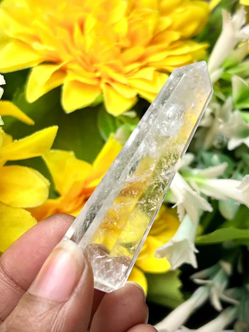 May include: A clear quartz crystal point, held against a backdrop of yellow and white flowers. The crystal is transparent with internal fractures and a pointed tip, showcasing natural formations. The background features bright yellow blooms and green foliage.