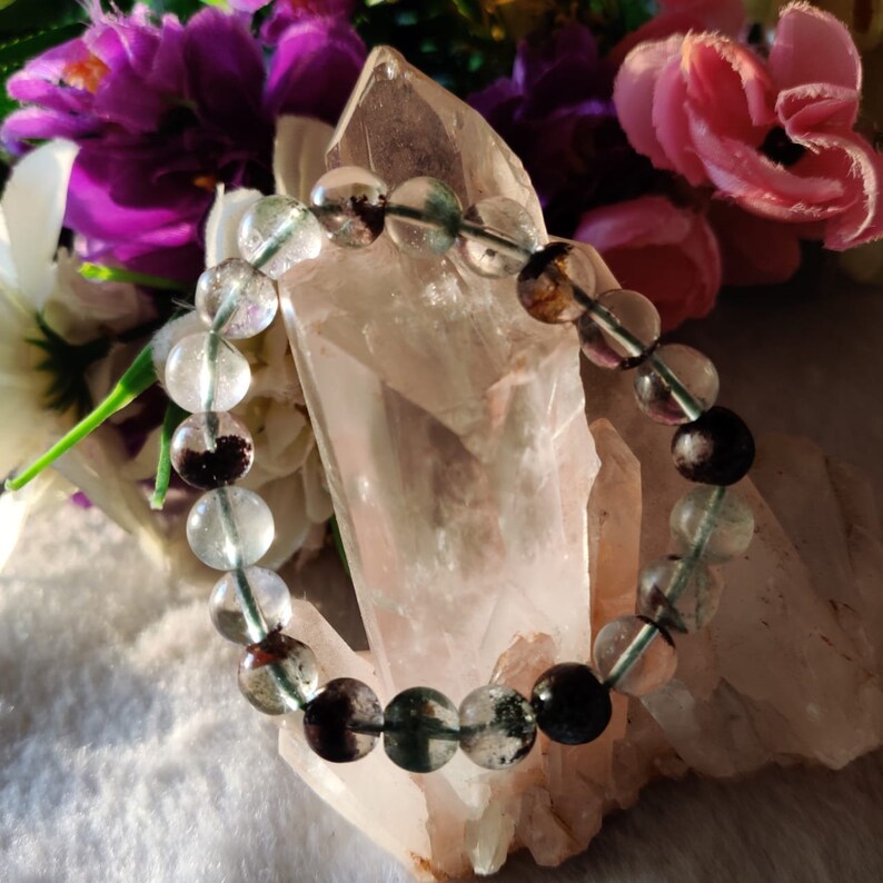 May include: A green and black gemstone bracelet with clear beads. The bracelet is displayed on a white crystal.