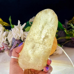 Puede incluir: Un objeto translúcido, de color amarillo pálido, alargado y redondeado, similar a una piedra, con una superficie cristalina texturizada. El objeto tiene un aspecto suave y brillante, y se sostiene en la mano, mostrando su forma orgánica irregular.