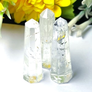 May include: Three clear quartz crystal points with hexagonal bases. The crystals are transparent with internal inclusions and a slight yellow hue at the base. The background features yellow flowers and white foliage.