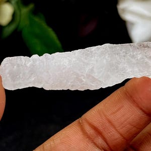 May include: A translucent, crystalline mineral specimen, held between fingers. The rough, elongated shape has a textured surface. The background is dark, with hints of green foliage and white flowers. This item is likely a raw crystal or gemstone.