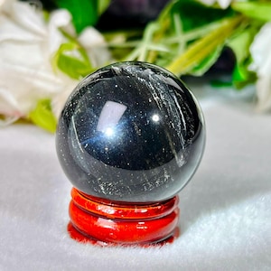Può includere: Una sfera nera lucida con sottili inclusioni argentate e una superficie riflettente, appoggiata su un supporto circolare in legno rosso lucido. La sfera presenta una forma liscia e arrotondata con riflessi di luce visibili sulla superficie.