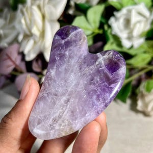 Peut inclure: Un outil de gua sha en forme de cœur taillé dans une pierre d'améthyste polie. La surface présente des variations naturelles de couleur, allant du violet profond au lavande pâle translucide, avec des fractures cristallines internes visibles.