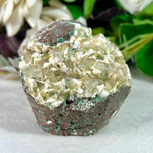 May include: A raw mineral specimen featuring a cluster of translucent, pale green prismatic crystals embedded in a dark brown, porous matrix. The crystalline structure displays sharp, angular facets with white mineral deposits throughout.