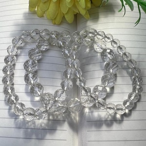 Pode incluir: Três fios de contas de cristal transparente, cada fio é um bracelete, com contas facetadas. Os braceletes estão sobre uma superfície branca riscada.