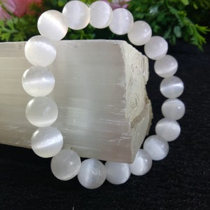 May include: White moonstone beaded bracelet with a smooth, polished finish. The bracelet is made of round beads and is displayed on a white selenite slab.