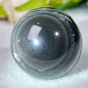 May include: A polished, spherical obsidian stone with a smooth, reflective surface. The dark, charcoal-grey material displays a subtle, shimmering metallic sheen across its rounded form, capturing light with a bright, white rectangular reflection.