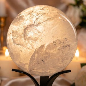 Op de afbeelding: Een heldere, bolvormige kristallen bol met zichtbare interne breuken en insluitingen, rustend op een zwarte metalen standaard. De bol straalt een warm, gloeiend licht uit dat de natuurlijke textuur en structuur van de steen benadrukt.