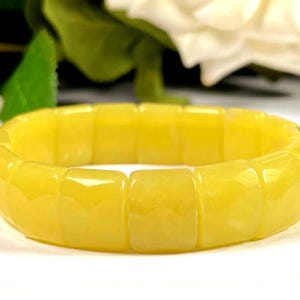 Peut inclure: Un bracelet jaune vif composé de pierres rectangulaires polies. Le bracelet est une bande de couleur unie, avec une surface lisse et réfléchissante. Le fond comprend des fleurs blanches et des feuilles vertes.