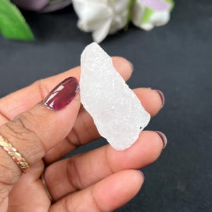 May include: A rough, white crystal with a textured surface. The crystal is held in a hand with maroon nail polish and a gold ring. The background is dark, with hints of white and green.