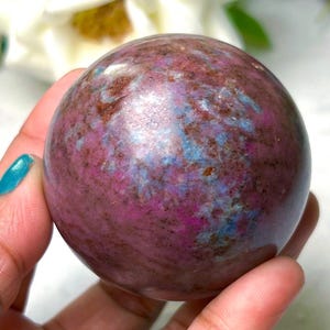 May include: A polished sphere with a mix of colors, including pink, brown, and blue. The gemstone ball is held in a hand, showcasing its smooth, rounded shape. The sphere is a decorative item.