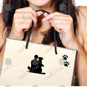 May include: A cream-colored shopping bag with black rope handles. The bag features a black silhouette of a person hugging a dog, along with two sets of black paw prints. The bag is held by a person.