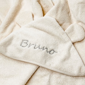 Puede incluir: Una toalla con capucha de color crema con el nombre "Bruno" bordado en gris en la capucha.