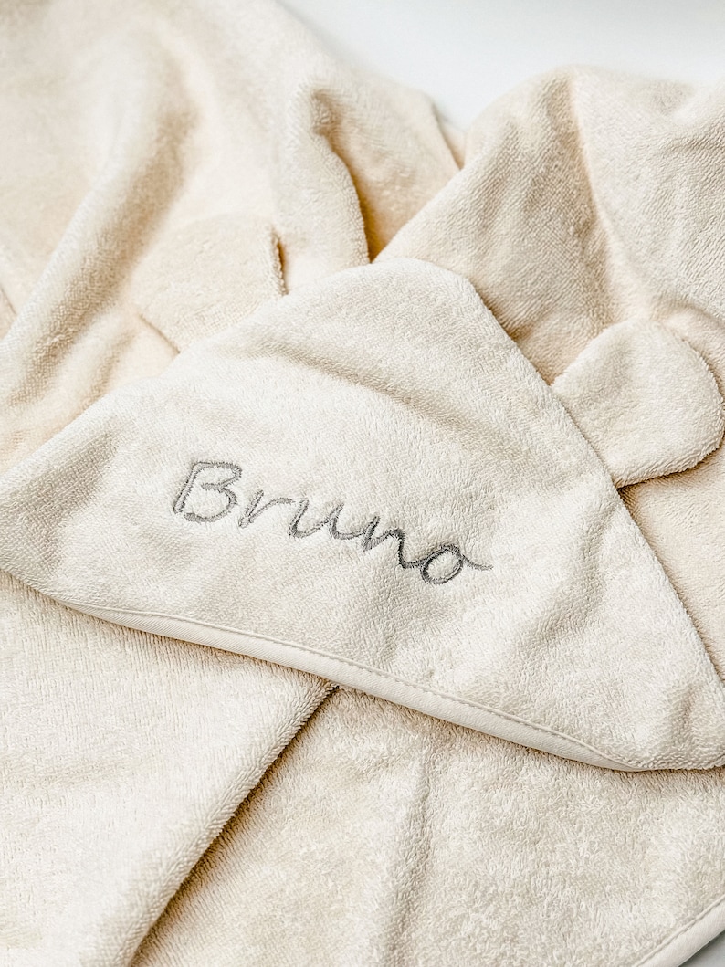 Puede incluir: Una toalla con capucha de color crema con el nombre "Bruno" bordado en gris en la capucha.