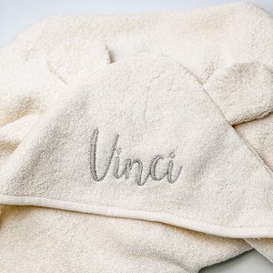 Puede incluir: Una toalla con capucha de color crema con el nombre "Vinci" bordado en gris en la parte delantera.