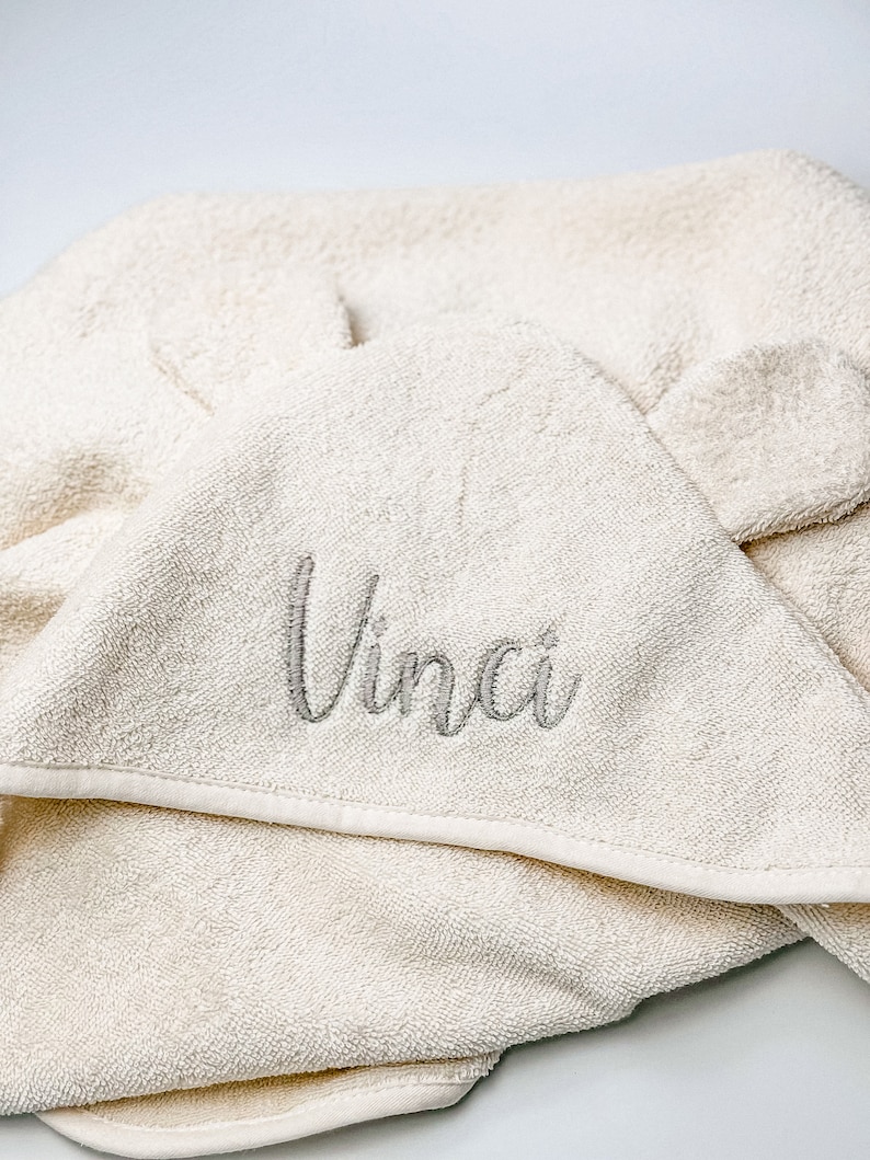 Puede incluir: Una toalla con capucha de color crema con el nombre "Vinci" bordado en gris en la parte delantera.
