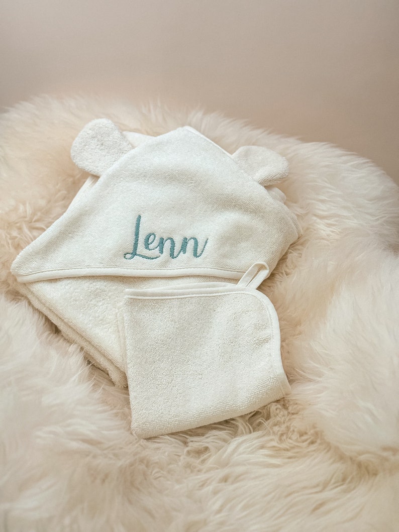Puede incluir: Toalla con capucha blanca con orejas de oso y un pa&ntilde;o de lavado blanco. La toalla tiene el nombre "Lenn" bordado en color turquesa.