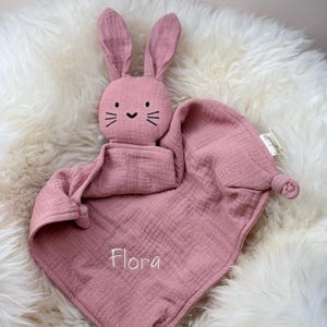 Könnte beinhalten: Eine rosa Hasen-Sicherheitsdecke mit dem Namen "Flora" bestickt. Die Decke ist aus weichem, strukturiertem Stoff und hat einen geknoteten Schwanz.