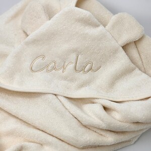 Puede incluir: Una toalla con capucha de color crema con el nombre "Carla" bordado en hilo dorado.