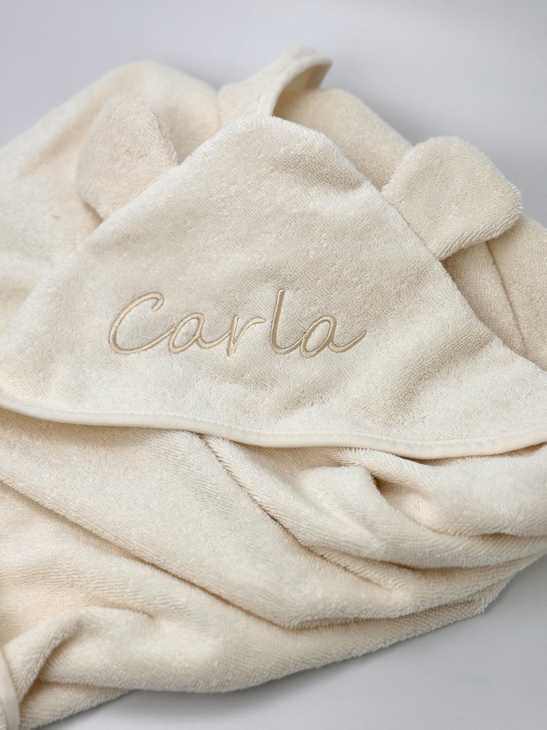 Puede incluir: Una toalla con capucha de color crema con el nombre "Carla" bordado en hilo dorado.